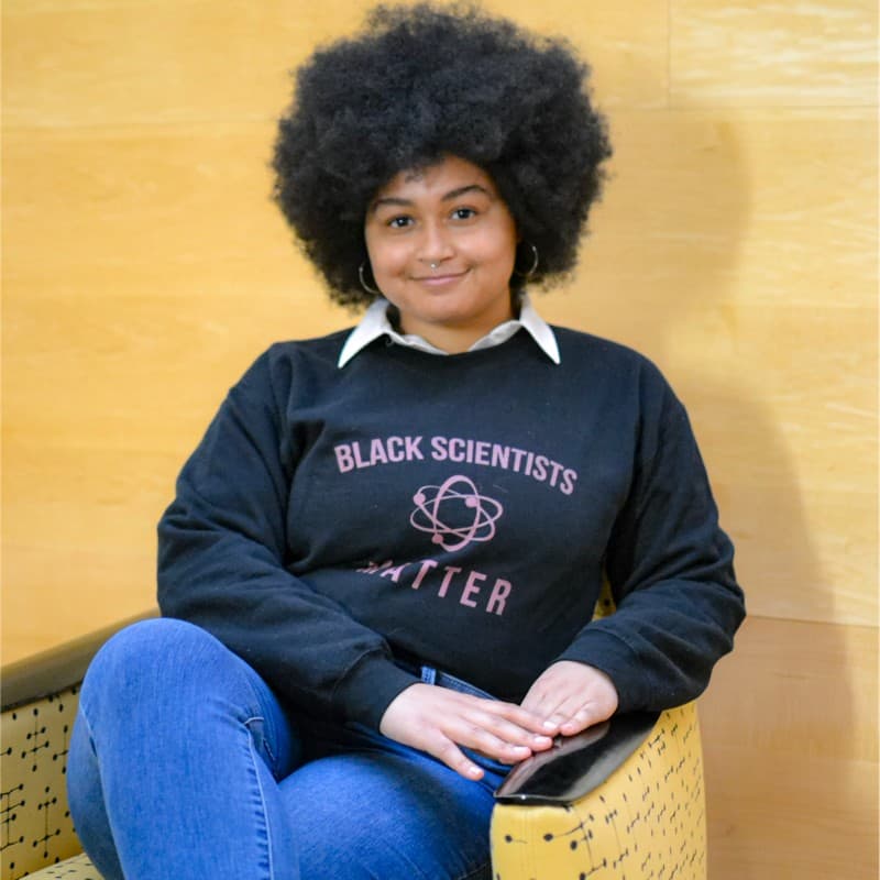 Kyla Smith sitting in a chair in front of a yellow wall smiling at the camera. Her black shirt reads "black scientists matter".